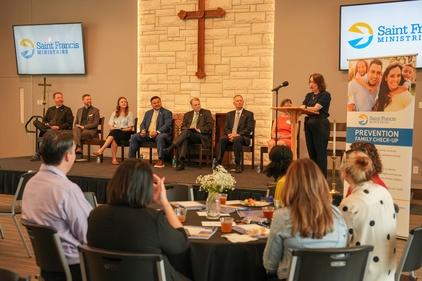 A group of seven people sit on a stage in panel discussion format. A woman stands at a podium speaking to an audience seated at round tables. Behind the stage is a stone wall with a cross and a Saint Francis Ministries logo. A banner promotes family check-ups.