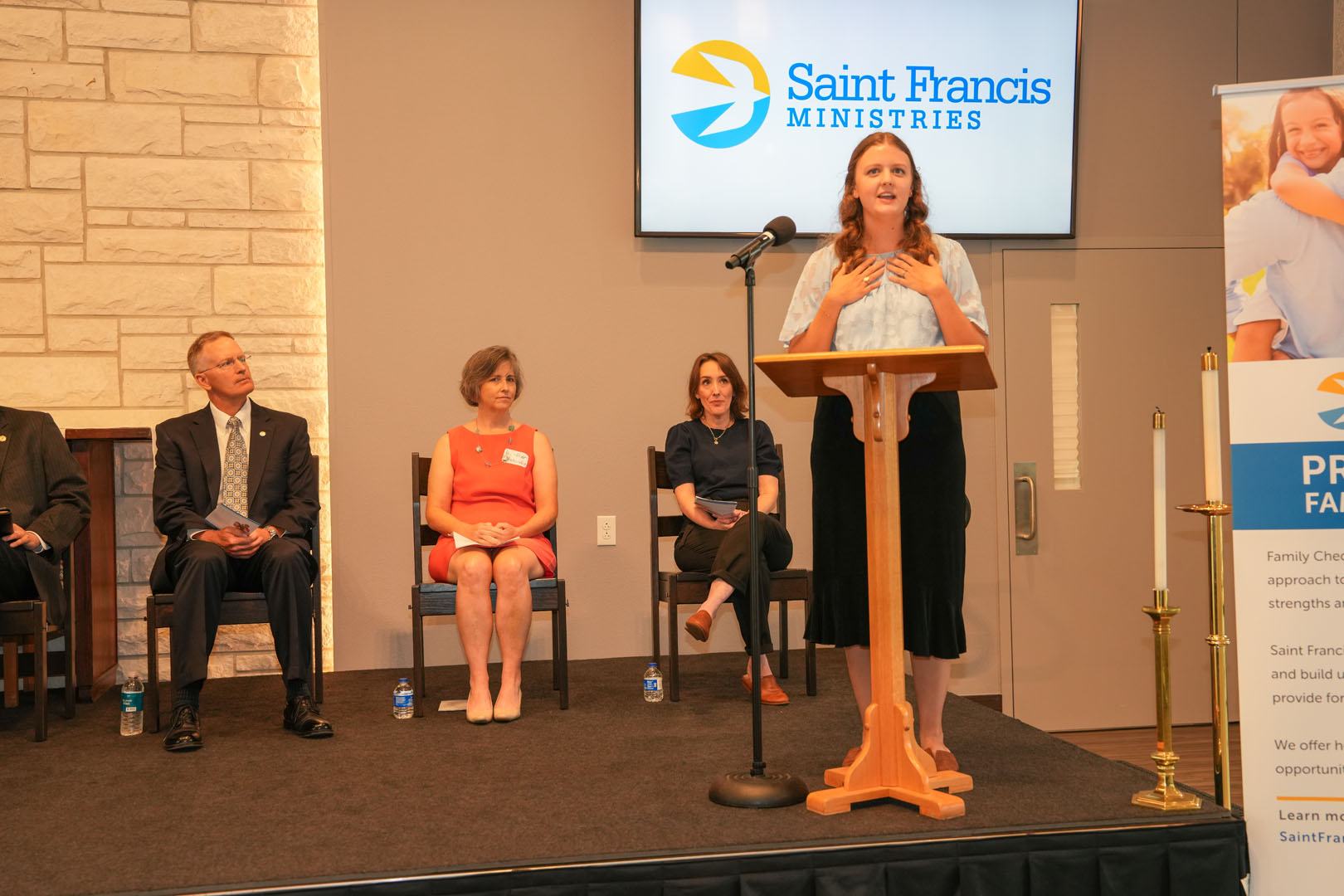 A woman is speaking at a podium under a sign that reads "Saint Francis Ministries." On stage, four people are seated, including a man and three women. The venue appears to be a conference or event space with a stone wall backdrop and a candle stand beside the podium.