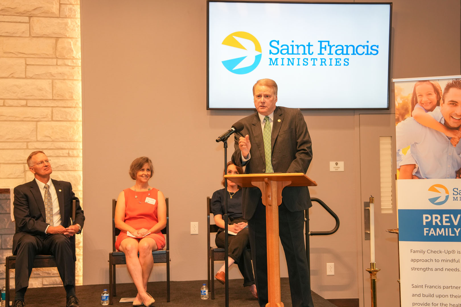 A man in a suit stands at a podium speaking, with three people seated behind him, two men and one woman. A large screen displaying the Saint Francis Ministries logo is visible on the wall behind them. A display board is also visible to the right.