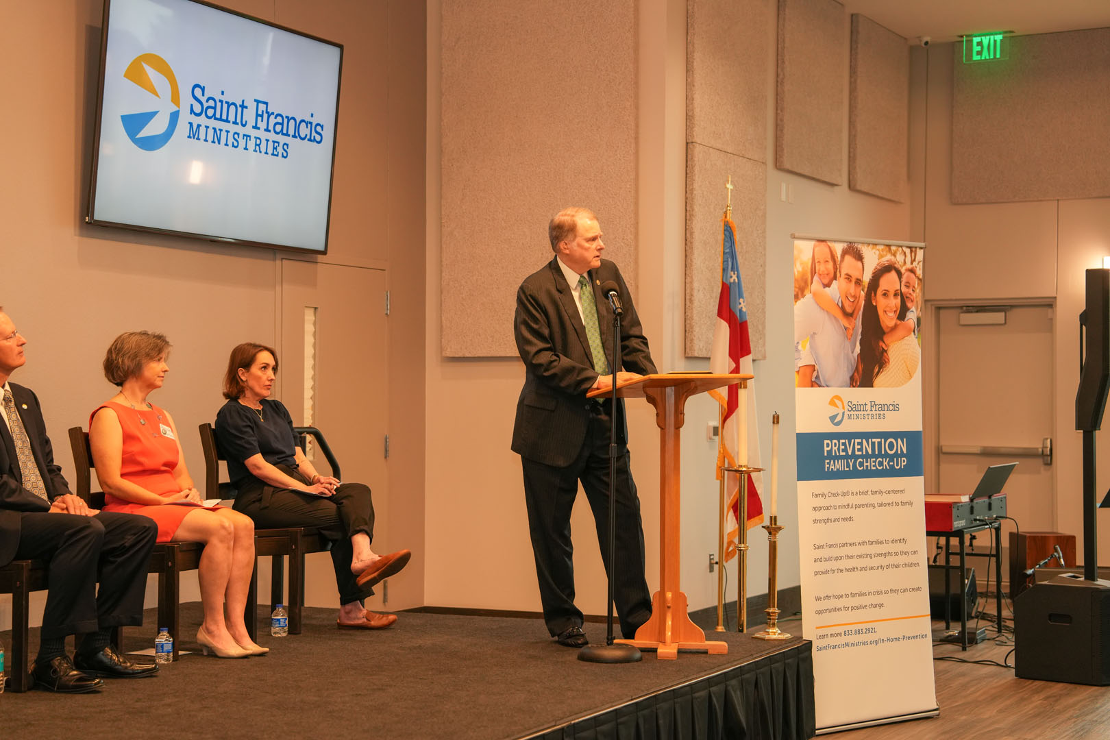 A man stands at a podium speaking into a microphone. Behind him on a screen is the logo for Saint Francis Ministries. Four people are seated on a stage to his left. A banner titled "Prevention Family Check-Up" is displayed near the podium.