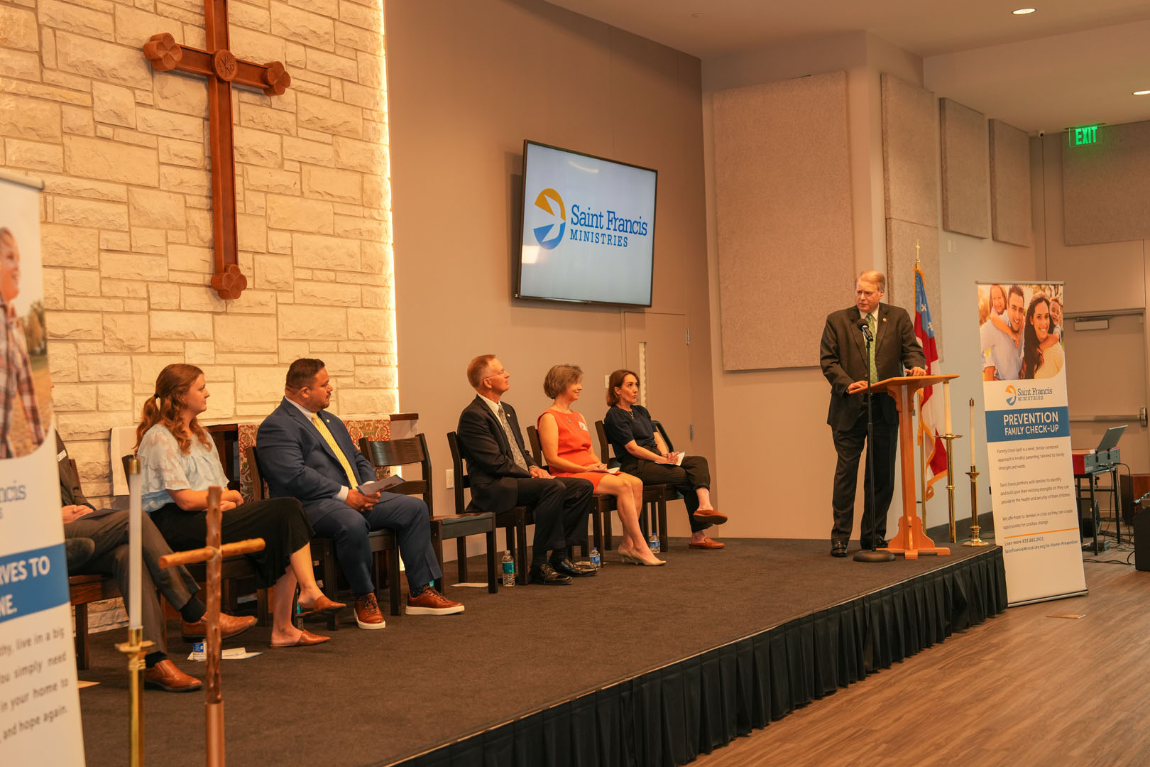 A man stands at a podium, speaking to an audience. Behind him, a banner for Saint Francis Ministries is displayed. Six people are seated on the stage, facing the audience. A large cross is mounted on the wall, and informational banners are visible on either side.