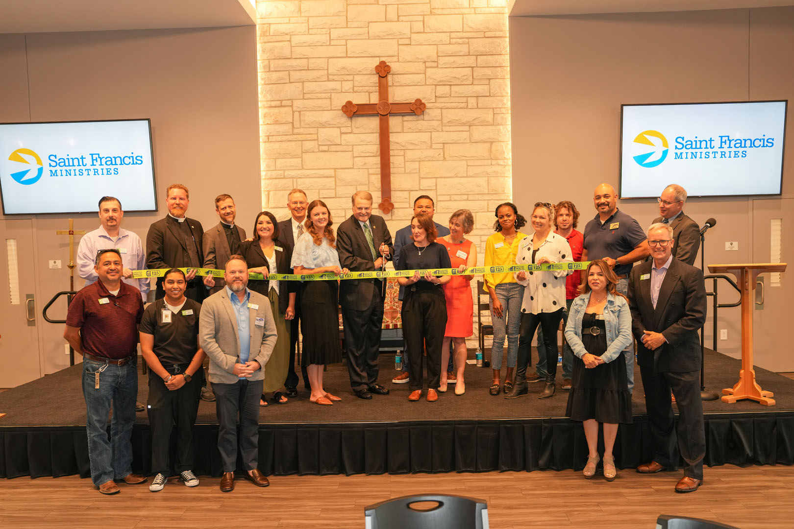 A group of people stands together on a stage for a ribbon-cutting ceremony, with a large pair of scissors held by the person in the center, in front of a cross. They are under a stone wall where a cross is mounted, and two monitors display "Saint Francis Ministries".