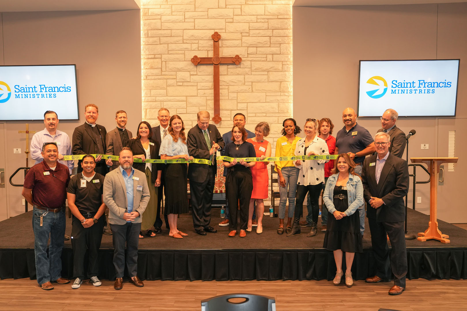 A group of people is standing in front of a table with a ribbon stretched out for a ribbon-cutting ceremony. There is a large wooden cross on the wall behind them and two screens displaying the "Saint Francis Ministries" logo.