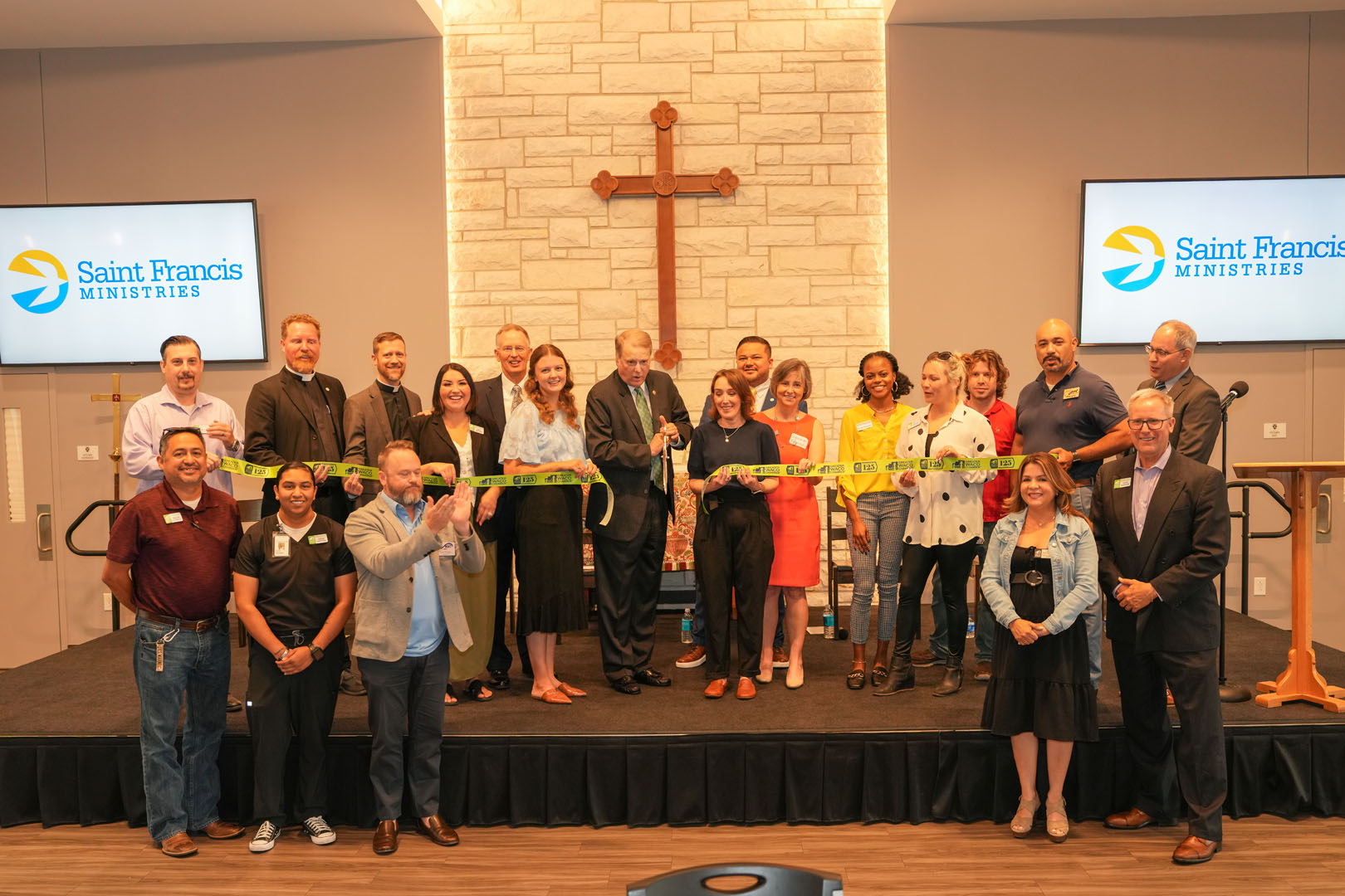 A group of people is gathered on a stage for a ribbon-cutting ceremony, with a large cross on the wall behind them. Two people in the center hold scissors to cut a ribbon. "Saint Francis Ministries" is displayed on two screens on either side of the stage.