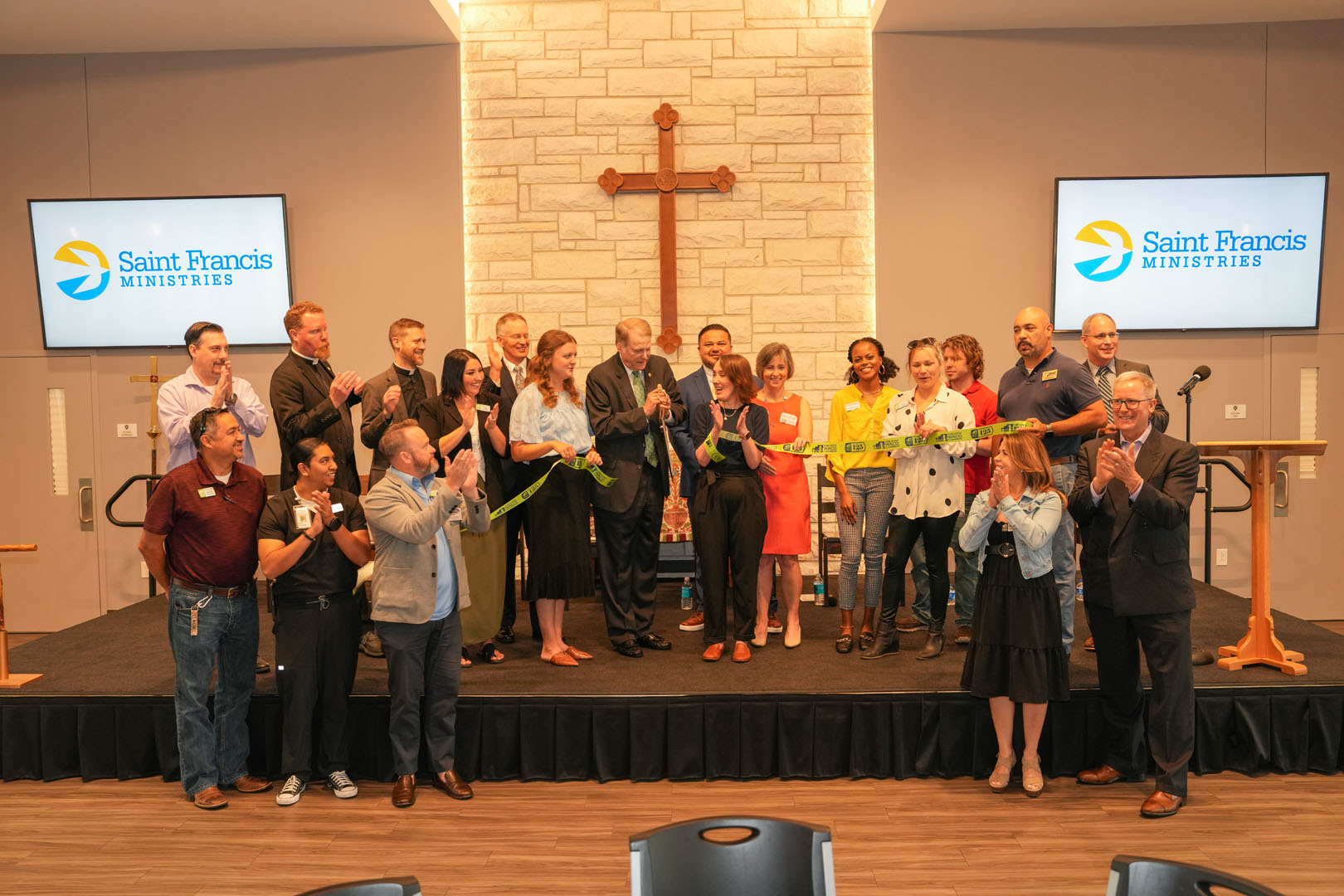 A large group of people stands on a stage with two screens displaying the Saint Francis Ministries logo. A ribbon is being cut in the center of the group, with everyone clapping or smiling. A large wooden cross is mounted on a stone wall in the background.