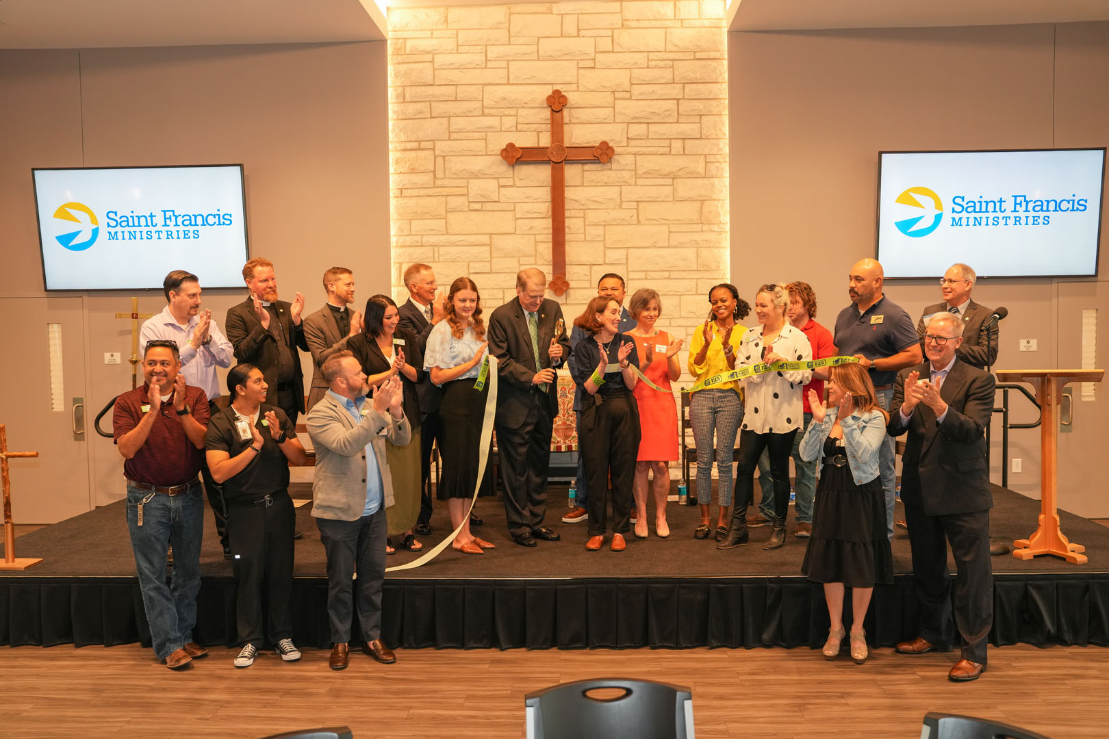 A group of individuals stands on a stage in front of a wall with a cross, with a few people cutting a green ribbon. Two screens display the "Saint Francis Ministries" logo. The group appears to be celebrating an event. Some are clapping and smiling.