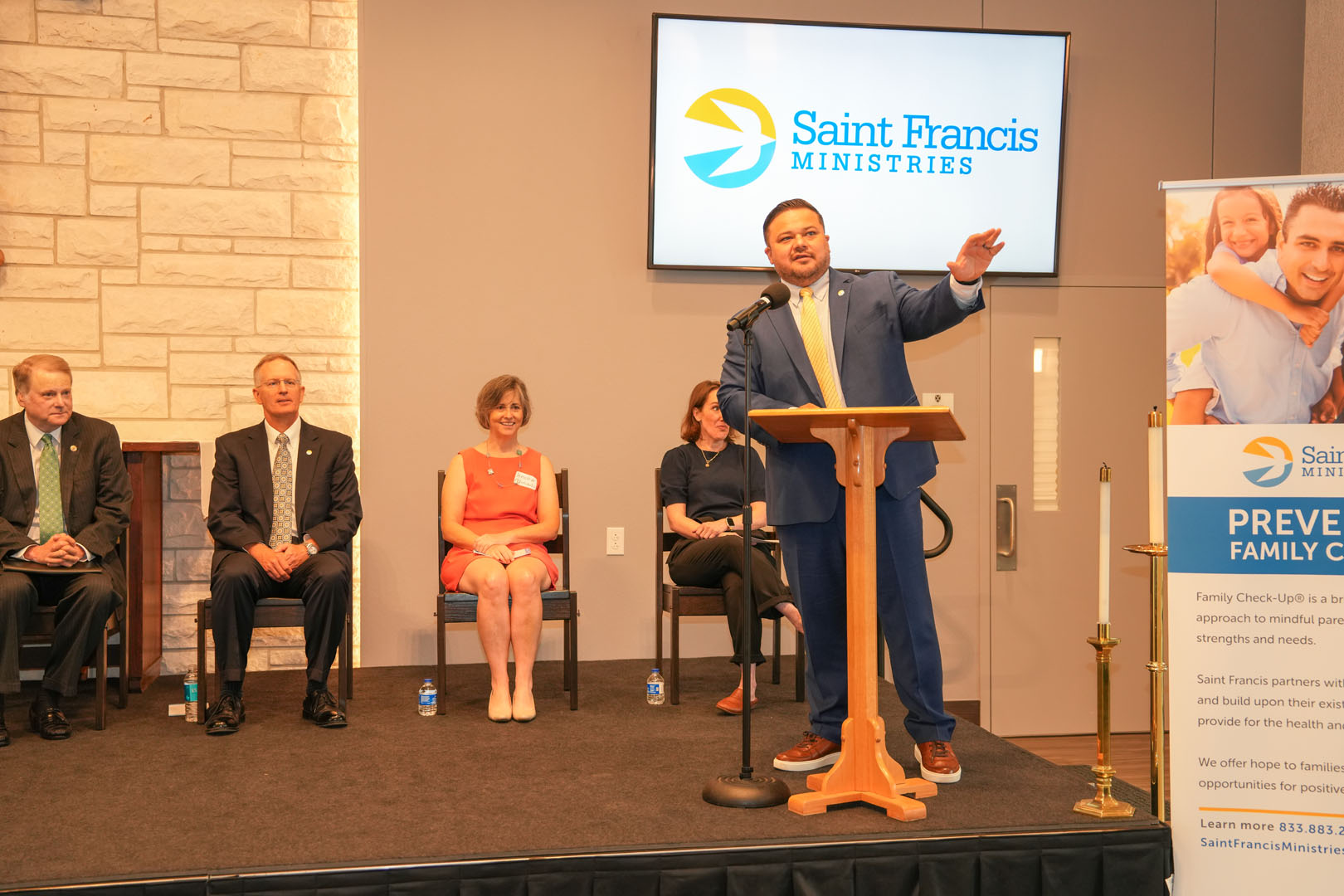 A man in a blue suit stands at a podium speaking to an audience at an event hosted by Saint Francis Ministries. Four people are seated behind him, and a banner with information about family check-ups is displayed nearby. A large logo is shown on a screen in the background.