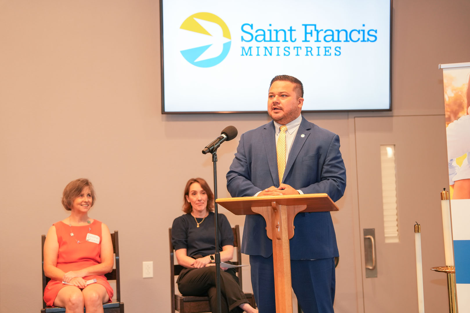 A man in a blue suit stands at a podium speaking into a microphone with the Saint Francis Ministries logo on a screen behind him. Two women sit in chairs to his left, one in a red dress with a nametag and the other in black.