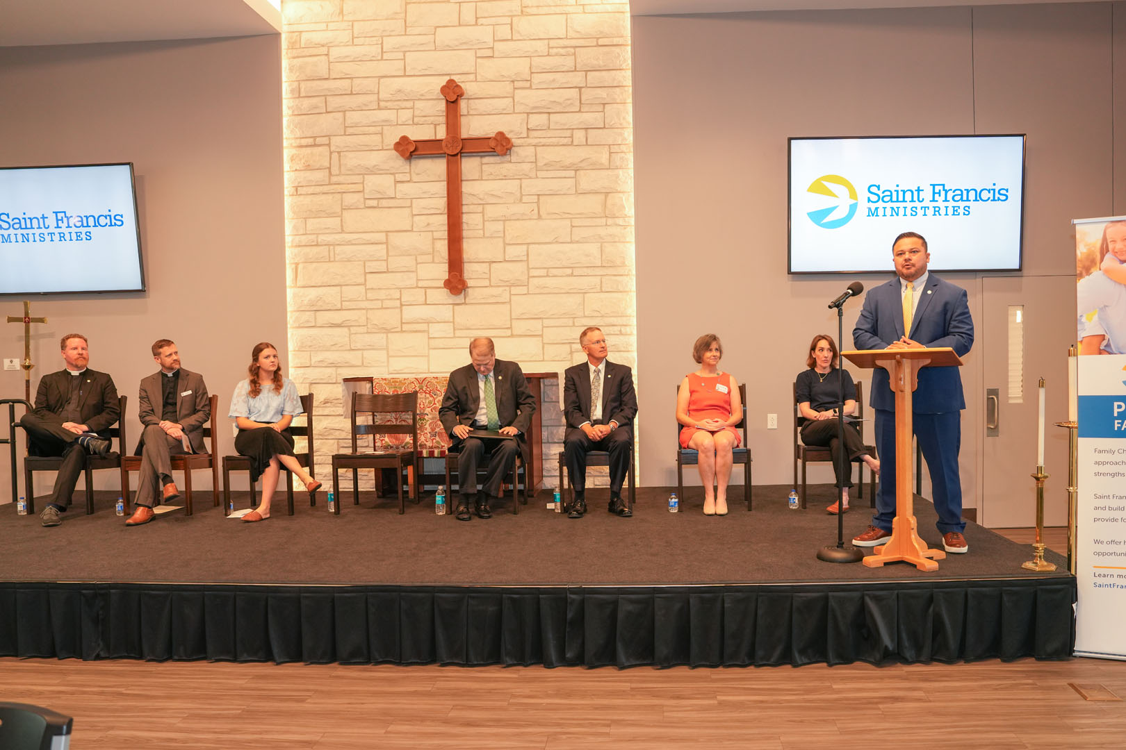 A group of seven people sits on a stage underneath a large wooden cross. A man in a suit stands at a podium speaking. Behind them, two monitors display the Saint Francis Ministries logo. To the right, a tall banner with text is partially visible.