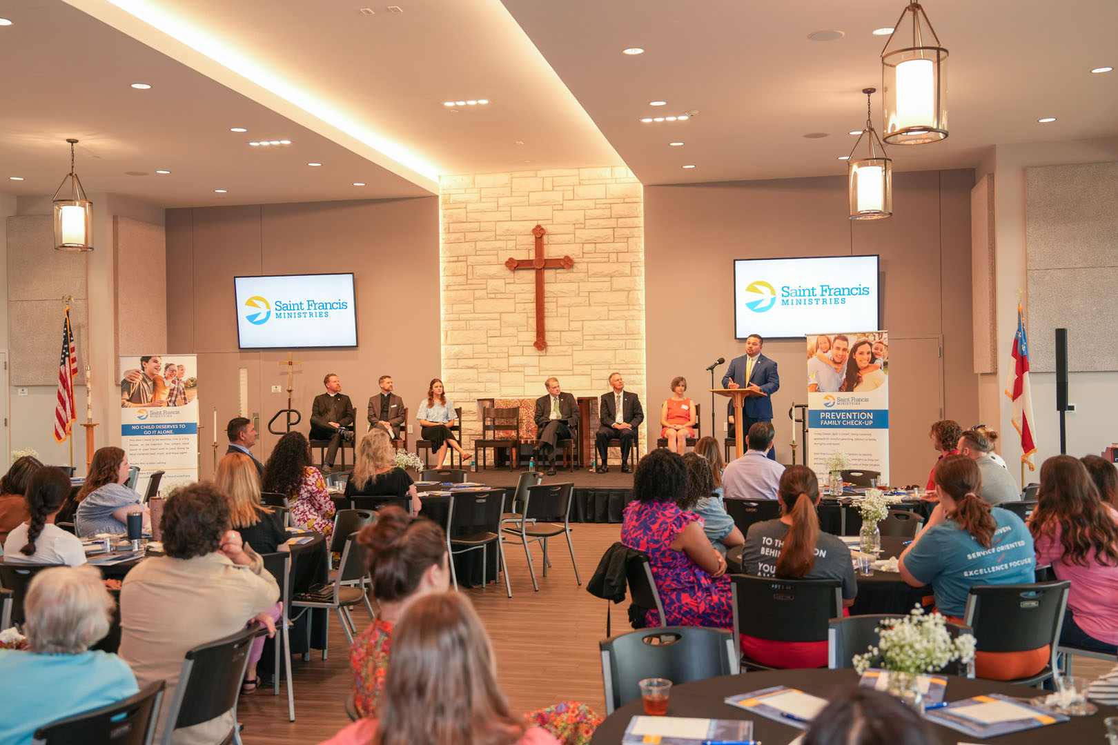 A conference room with chairs arranged in rows, facing a stage with seven people seated and one person standing at a podium. The backdrop features a stone wall with a cross. Two screens display the Saint Francis Ministries logo. Attendees sit at tables.