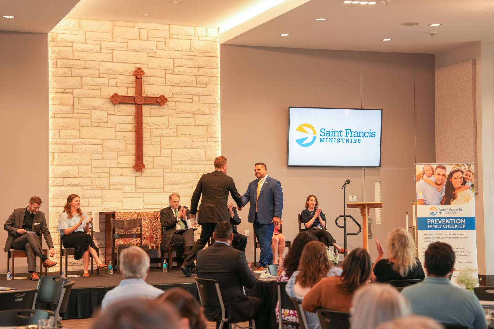 A man in a suit stands on a stage shaking hands with another man at a podium in front of a large screen displaying the Saint Francis Ministries logo. Several people are seated on the stage, and an audience is visible in the foreground.