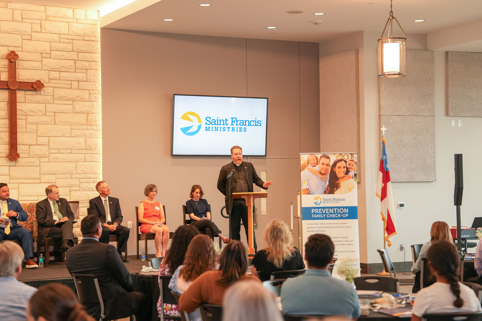 A person speaks at a podium during an event at Saint Francis Ministries. Attendees are seated facing the podium. Several people sit on chairs at the left. A poster features a "Prevention" slogan, and a wooden cross is mounted on the wall.