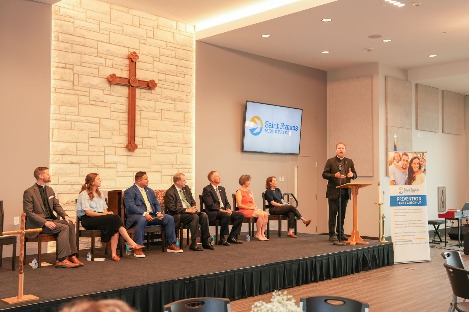 A group of eight people, dressed in business attire, sit on a stage with a speaker at a podium addressing the audience. A Saint Francis Ministries banner and a large cross on a stone wall are visible in the background.