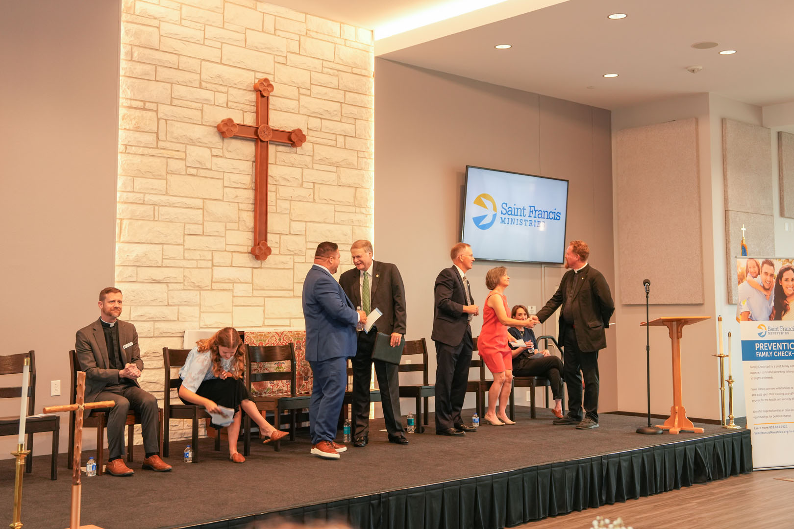 A group of people is standing on a stage near a podium. There is a large wooden cross on the wall behind them. A banner with the text "Saint Francis Ministries" is also visible on the stage. Some individuals are seated, and one person is leaning down.