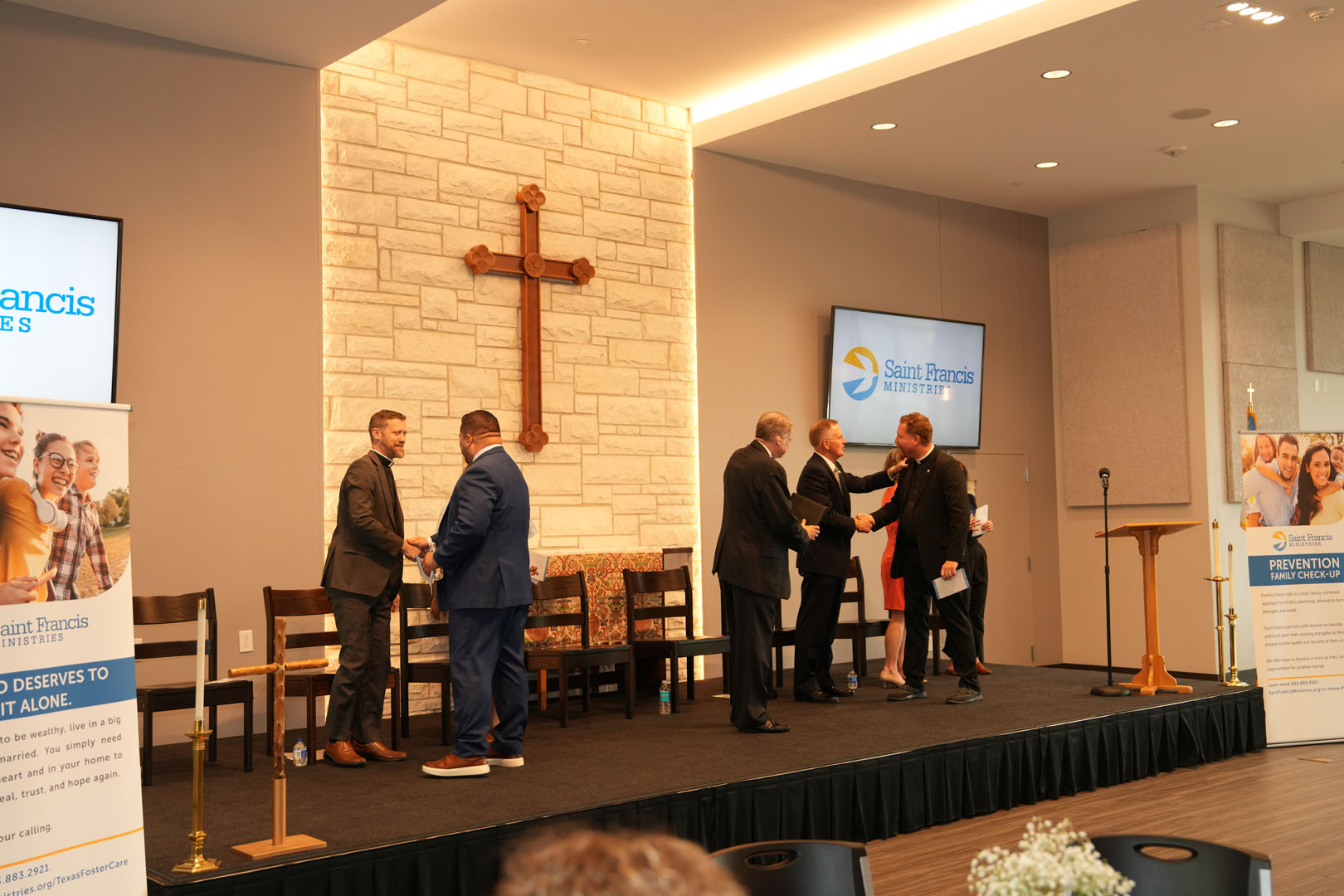 A group of people stands on a stage in a church-like setting with a large cross on the wall behind them. Several men in suits are shaking hands while others wait. Banners for Saint Francis Ministries are displayed on either side of the stage.