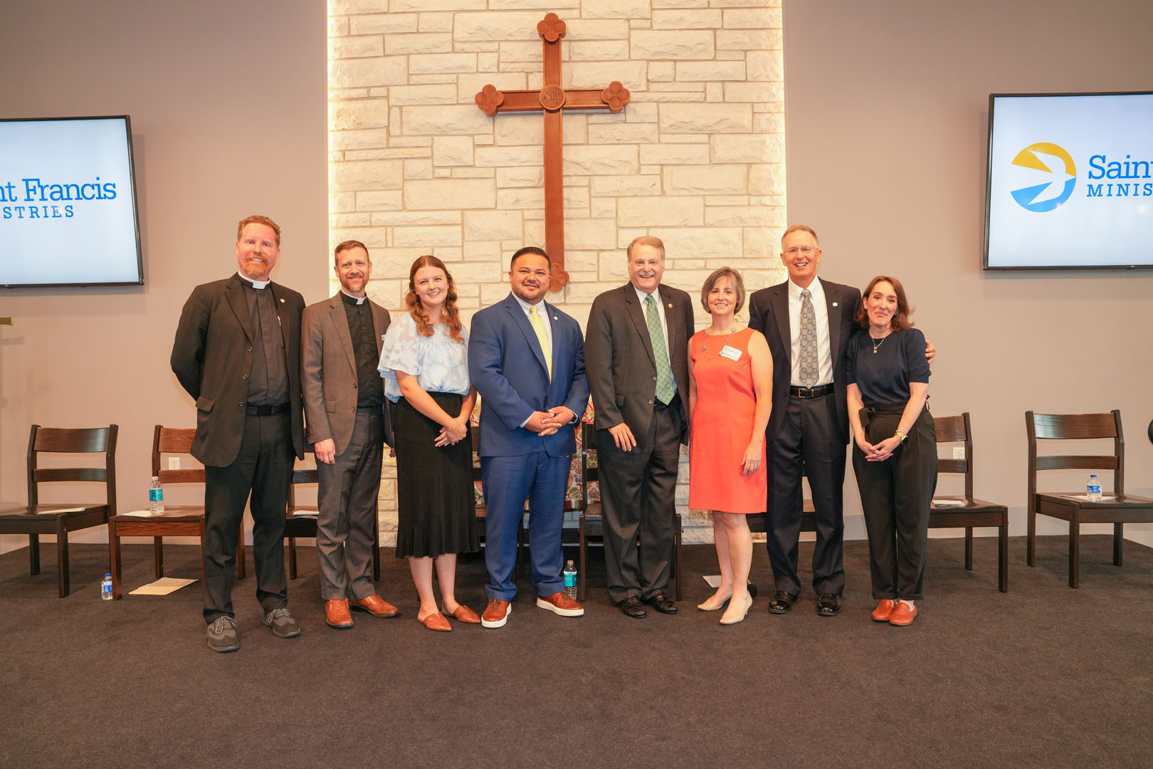 A group of nine people, consisting of six men and three women, stand in a line in front of a stone wall with a large wooden cross. They are in a room with chairs, a floor mat, and two screens displaying the Saint Francis Ministries logo.