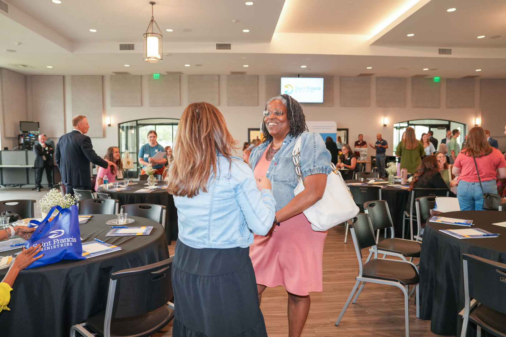 Two women engaged in conversation stand in the center of a room set up for a conference or event. They are surrounded by empty chairs and tables adorned with pamphlets and name cards. Other attendees are mingling in the background.