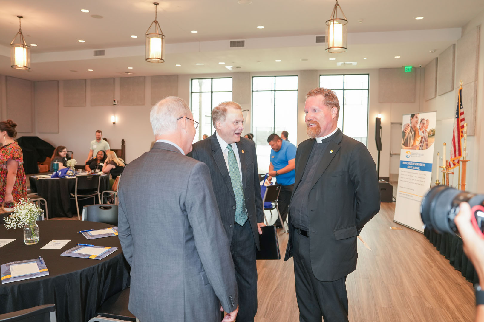 Three men in formal attire are conversing in a well-lit conference room. The room has round tables, informational brochures, and a banner display on the right side. Other attendees are seen mingling or seated in the background. An American flag stands in the corner.