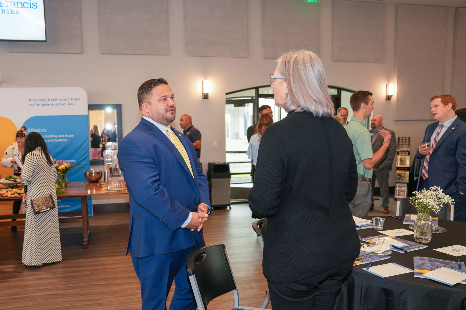 Several people are engaged in conversation in a well-lit room. A man in a blue suit talks to a woman with gray hair. A display table with informational materials and flowers is in the foreground. Additional individuals are interacting in the background.