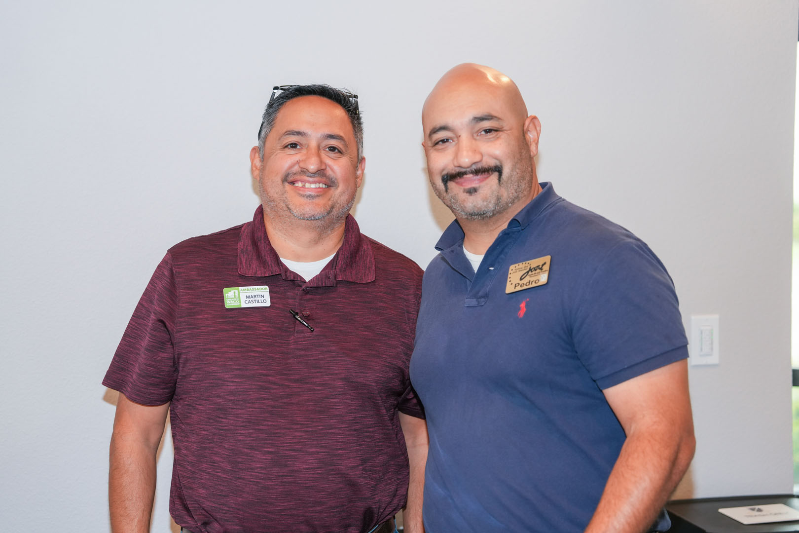 Two men are standing side by side and smiling. The man on the left is wearing a maroon polo shirt with a green and white name tag. The man on the right is wearing a blue polo shirt with a gold name tag. They are in a room with a white wall in the background.