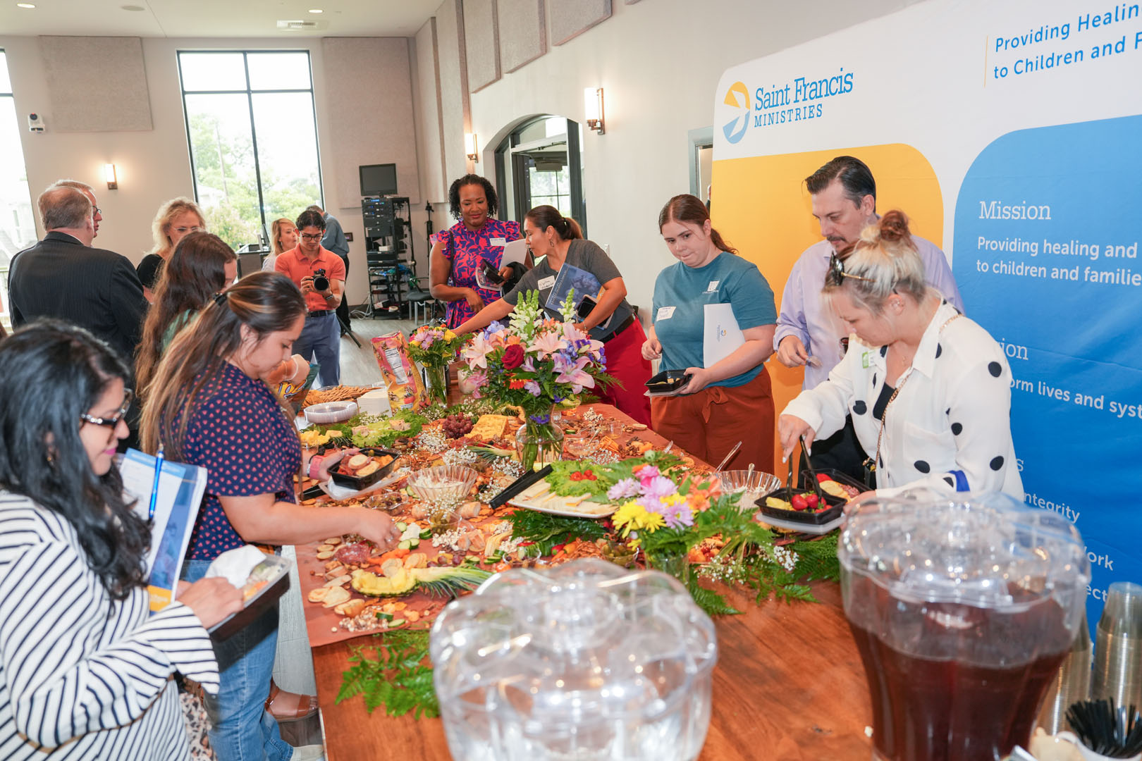 People are gathered around a table with a variety of food at a Saint Francis Ministries event. They are serving themselves from a buffet that includes fruits, cheeses, and other dishes. A large informational banner about Saint Francis Ministries is displayed in the background.