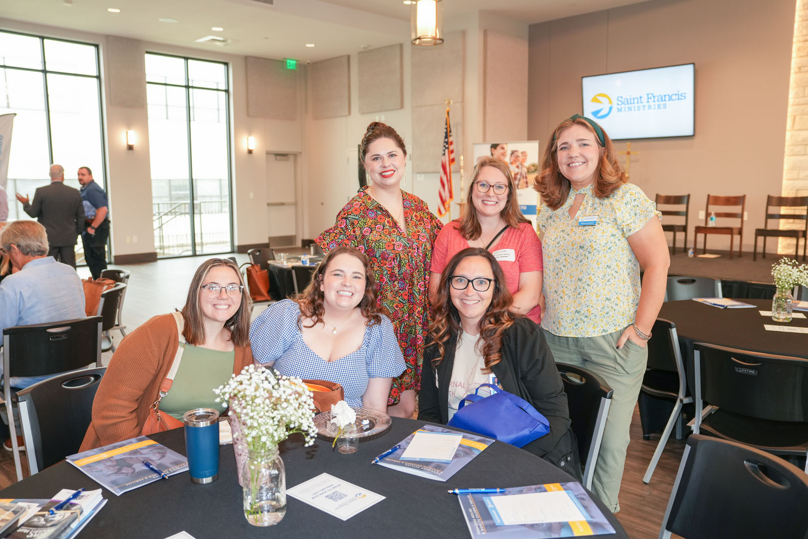 Six smiling individuals pose together around a table with event materials and floral centerpieces in a modern room. Behind them is a large screen displaying the logo "Saint Francis Ministries" and a United States flag.