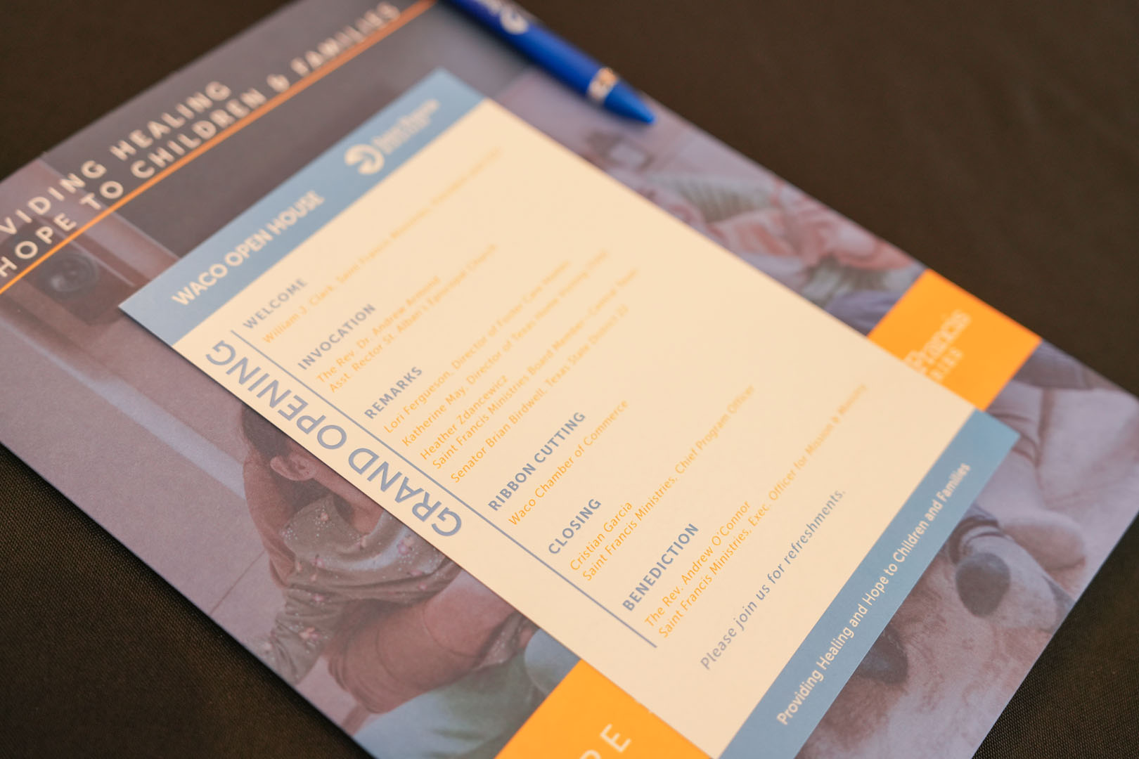 A printed event program titled "Waco Open House" is seen placed on a table, accompanied by a blue pen. The agenda includes welcome remarks, invocation, ribbon cutting, closing, and benediction. The program is for a grand opening and mentions refreshments are available.