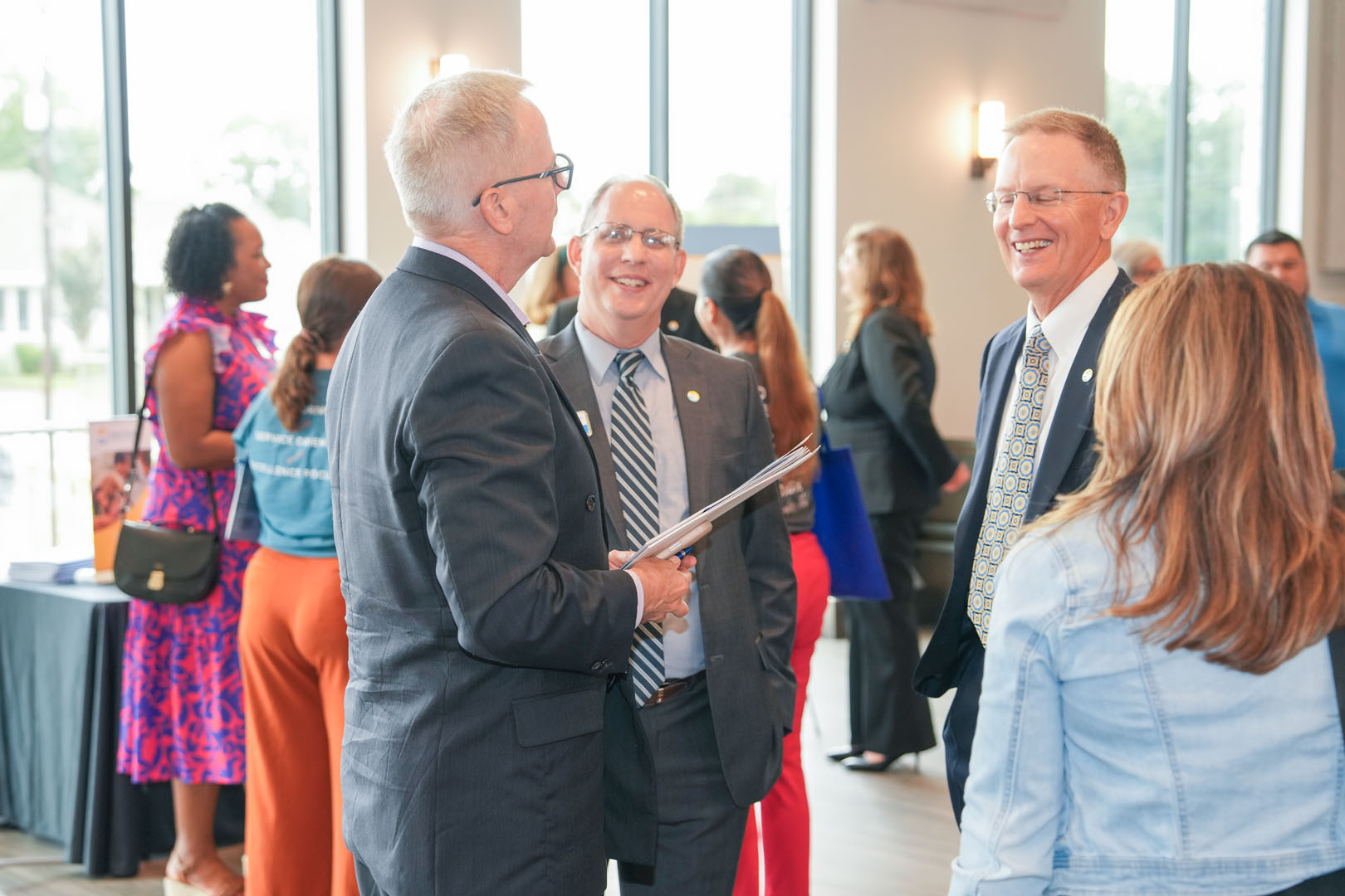 A group of people are attending a formal indoor event. Three men in suits are engaged in conversation and smiling, while others in the background are talking and mingling. The room is well-lit with large windows and modern decor.