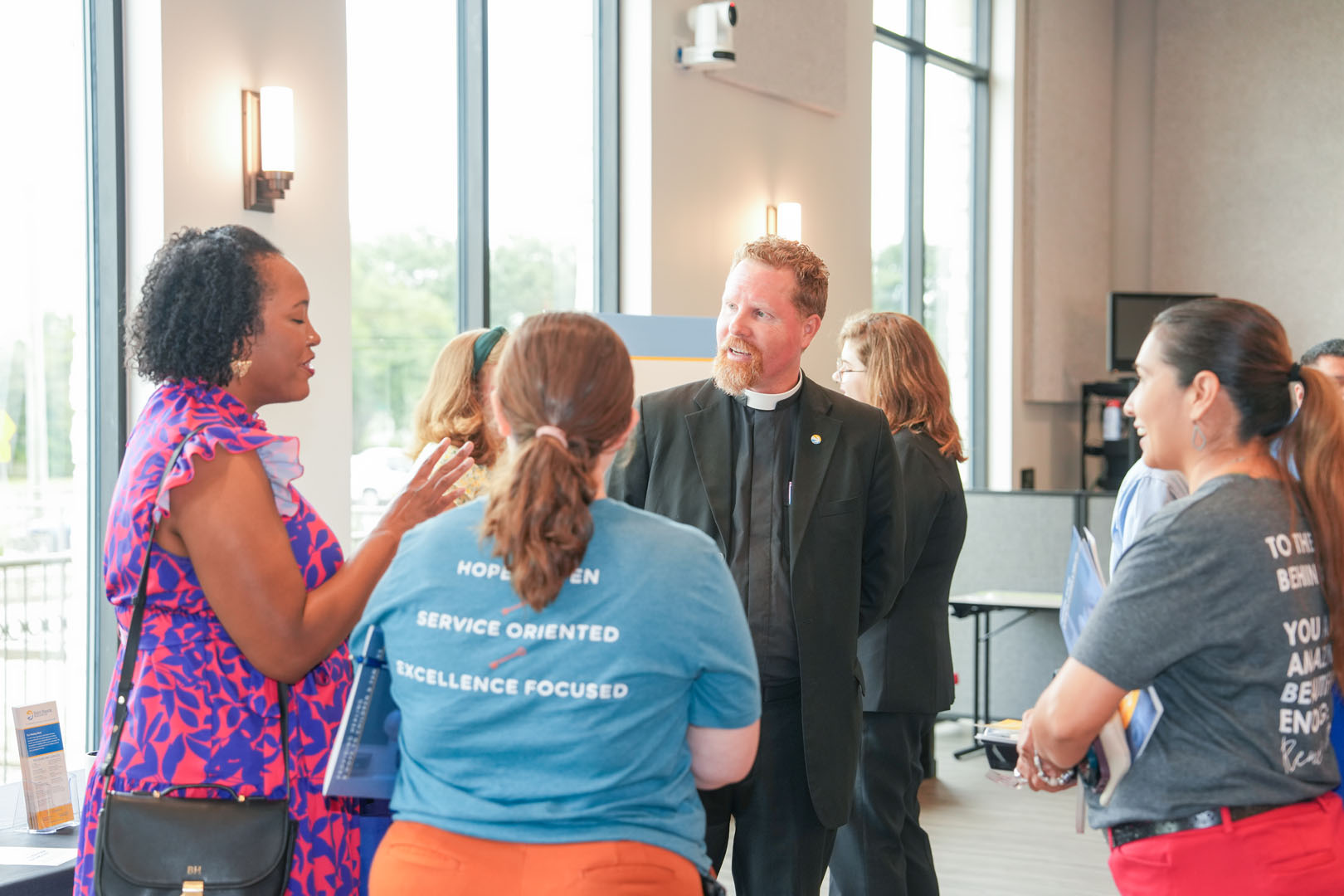 A group of people are engaged in conversation by large windows. A woman in a purple and pink dress talks to a man in a clerical collar and several other individuals facing each other. Light fixtures illuminate the room, and large windows overlook an outdoor scene.