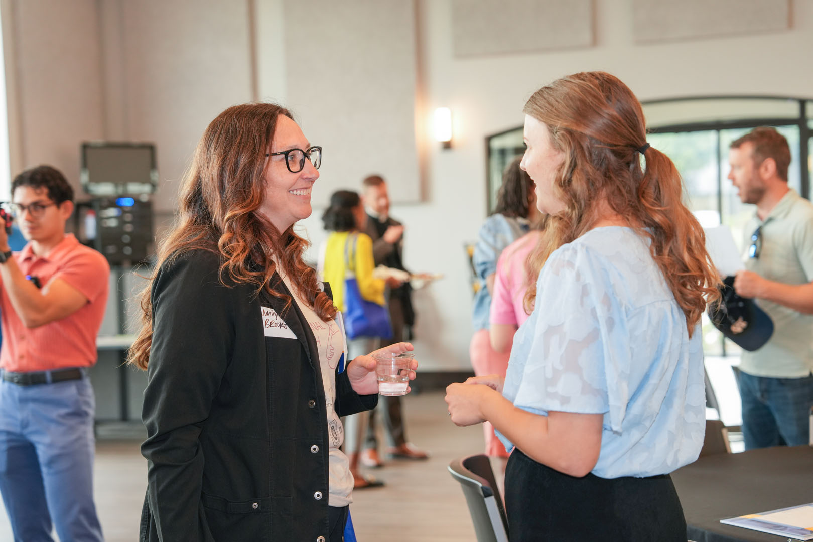 Two women are engaged in conversation at a casual indoor gathering. One is holding a glass of water and wearing a name tag. In the background, other people are mingling and talking. The room has large windows and a bright, airy feel.