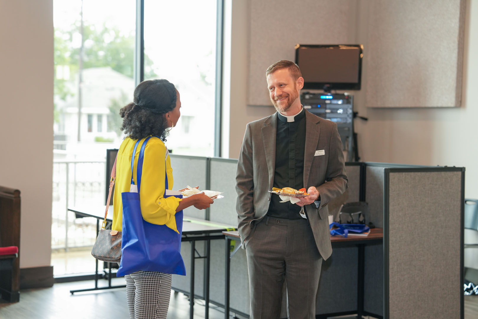 An individual in a yellow top and a blue bag talks with a religious figure in a suit and clerical collar. Both are holding plates of food and standing in a room with tables, chairs, and a window in the background. The setting looks like a casual event.