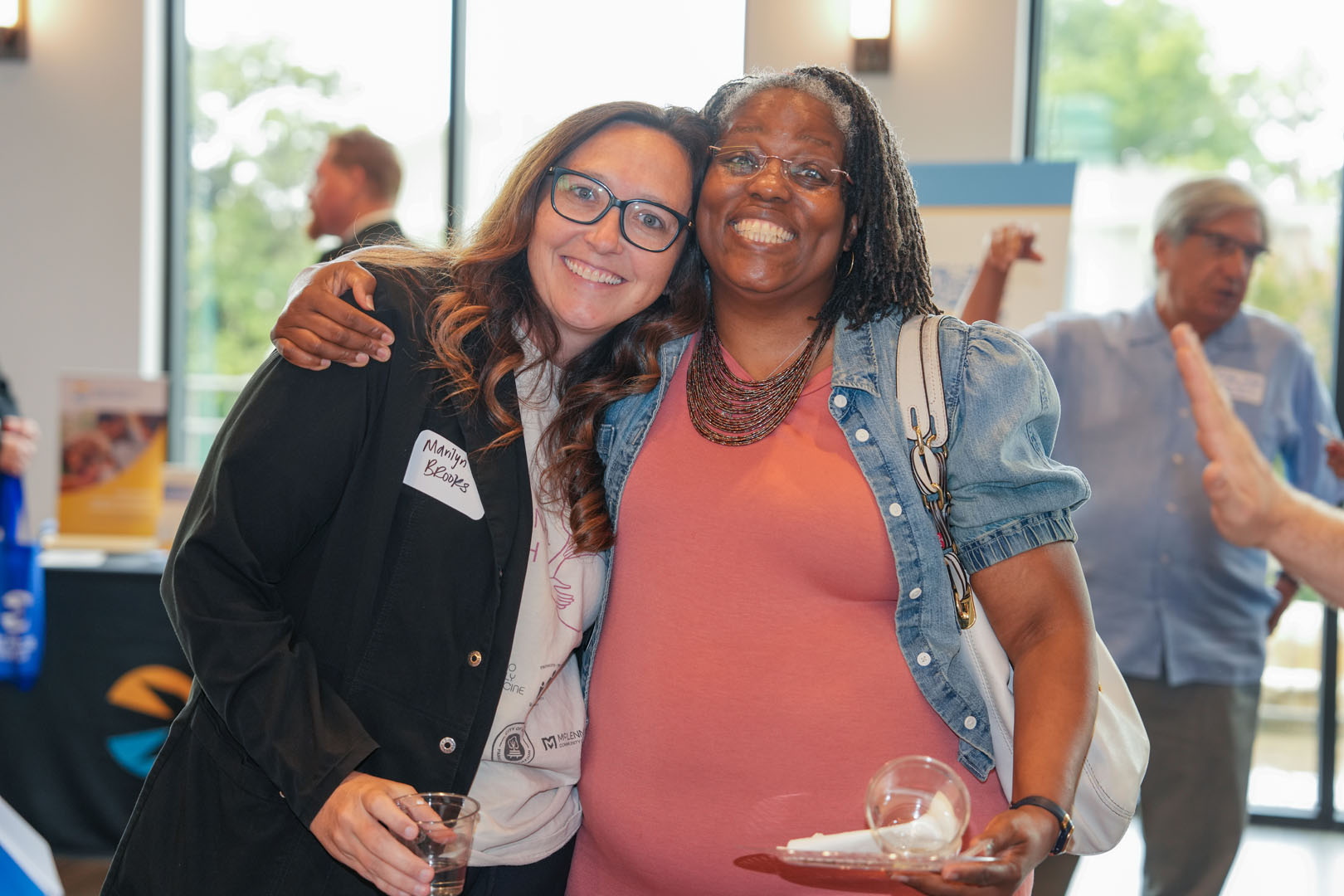 Two women are standing close together, smiling at the camera. The woman on the left has long hair and is holding a drink, while the woman on the right, wearing glasses and a red dress, carries a plate with a drink in one hand. Other people are in the background.