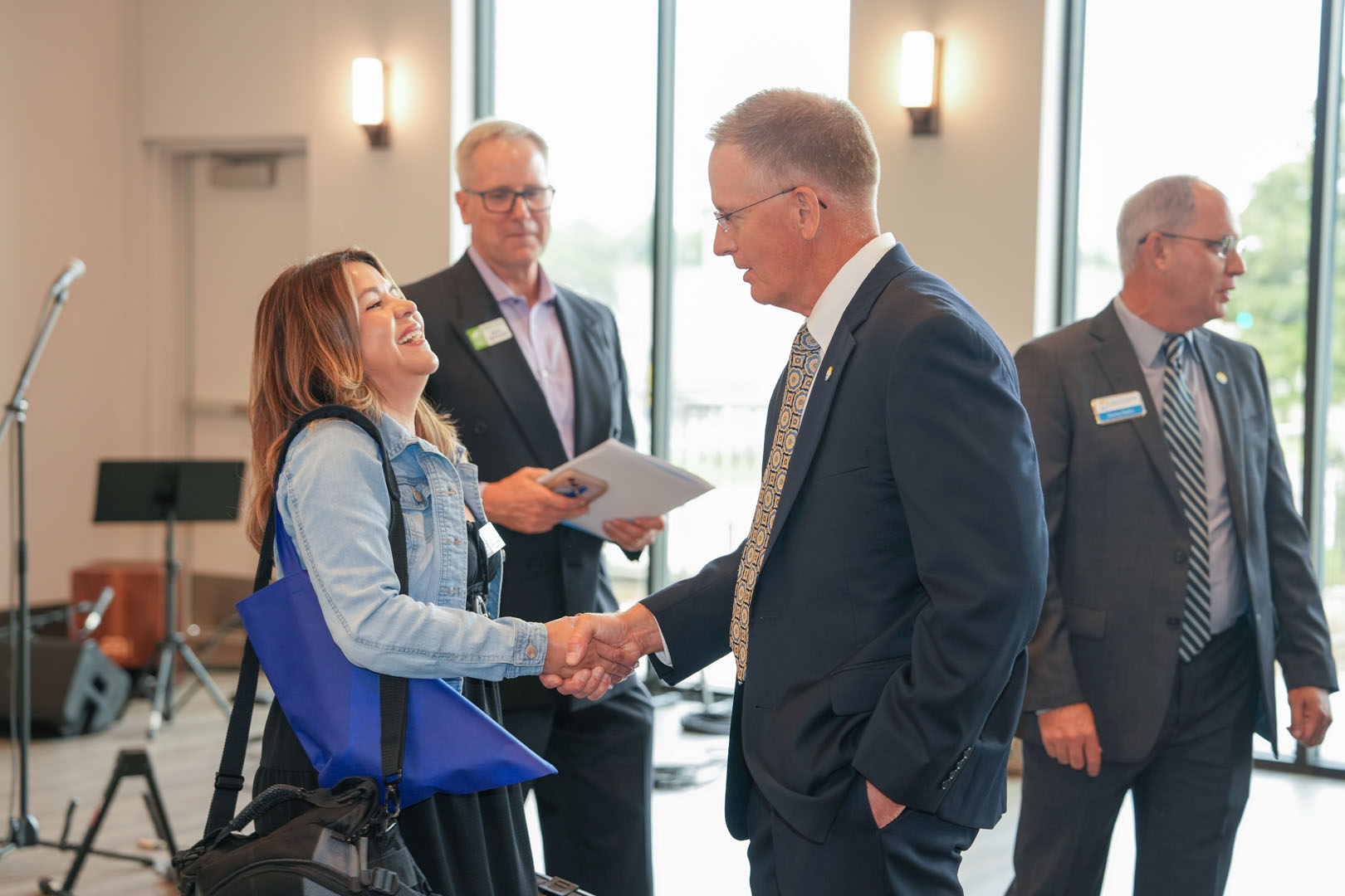 Four people are gathered in a room with large windows. A man in a suit is shaking hands with a woman carrying a blue bag and another bag. Two other men, also in suits, are standing in the background, holding papers and watching the interaction.