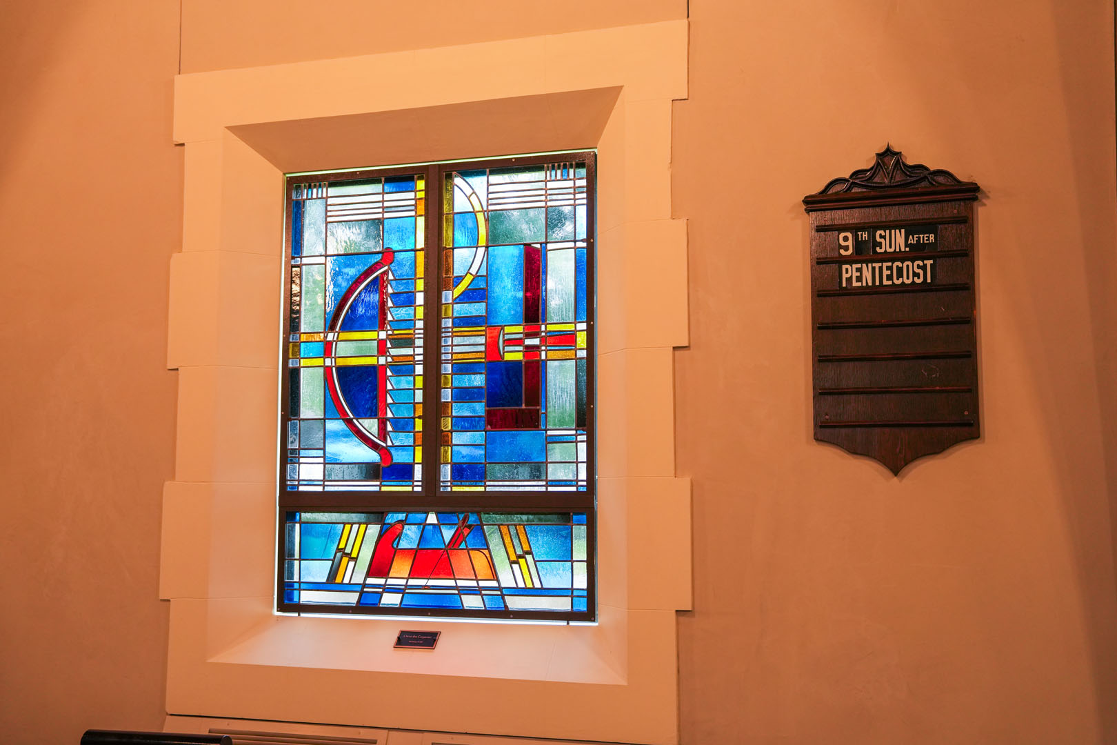 A stained glass window featuring abstract geometric designs and vibrant colors is set in a white frame on a wall. To the right, a wooden plaque reads "9th Sun After Pentecost." The room appears to be softly lit.