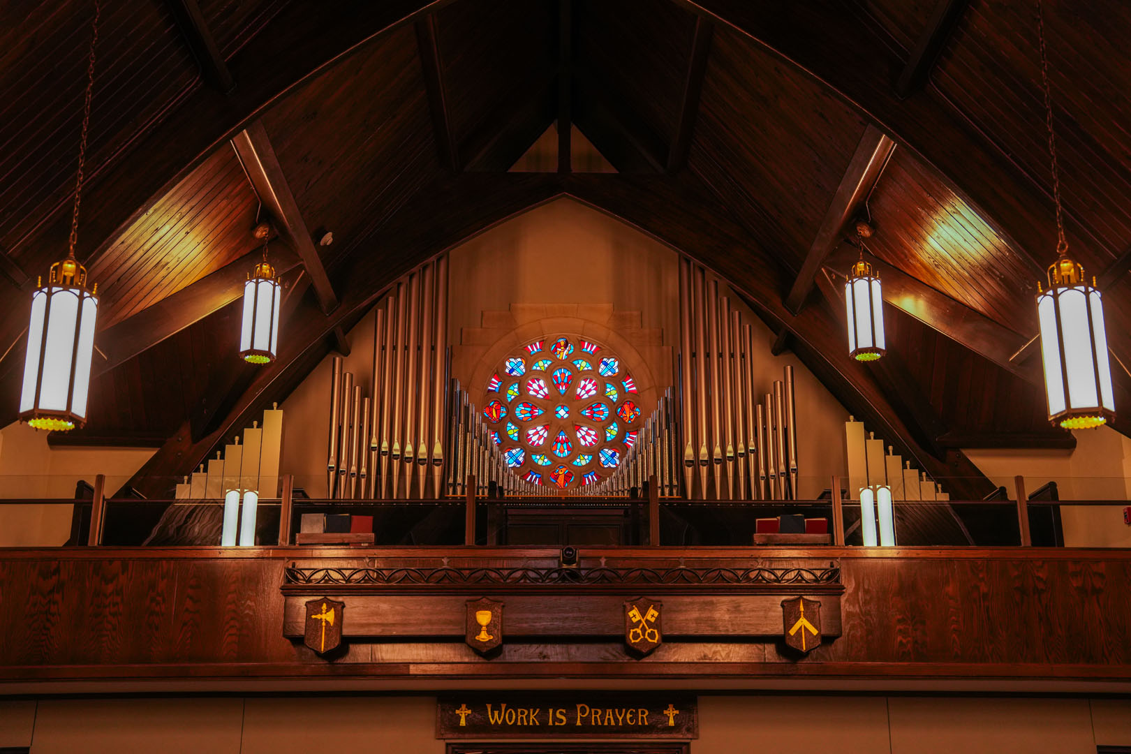 The image shows the interior of a church with a wooden ceiling. There is a large pipe organ and a rose-shaped stained glass window above it. The lights are on, and the text "WORK IS PRAYER" is visible on a wooden panel below the organ pipes.