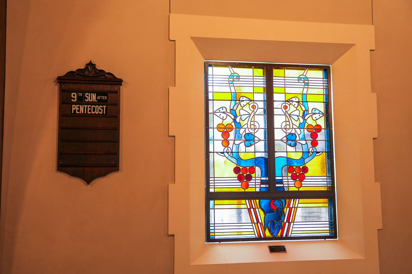 Stained glass window with a blue and yellow design featuring floral patterns. To the left, there is a mounted placard that reads "9th Sun. after Pentecost." The window is framed by a simple white wall.