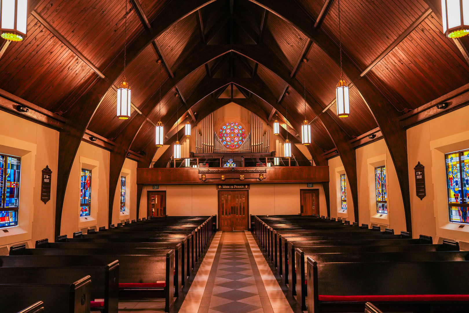 Interior of a church with wooden pews arranged in rows, a central aisle, and a high arched wooden ceiling. Stained glass windows line the walls and a large stained glass window is above the entrance. Modern hanging lights illuminate the space.