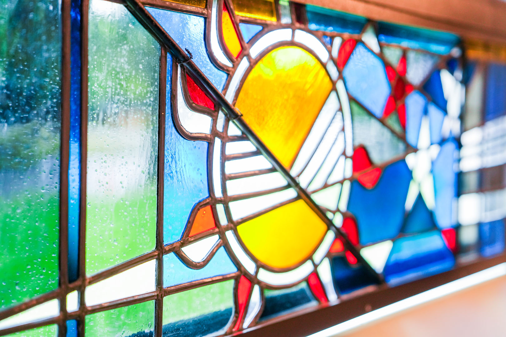 Stained glass window featuring a colorful design with a prominent central sun motif in yellow and orange. Surrounding elements include abstract shapes in blue, red, and green. Natural light filters through, illuminating the vibrant colors.