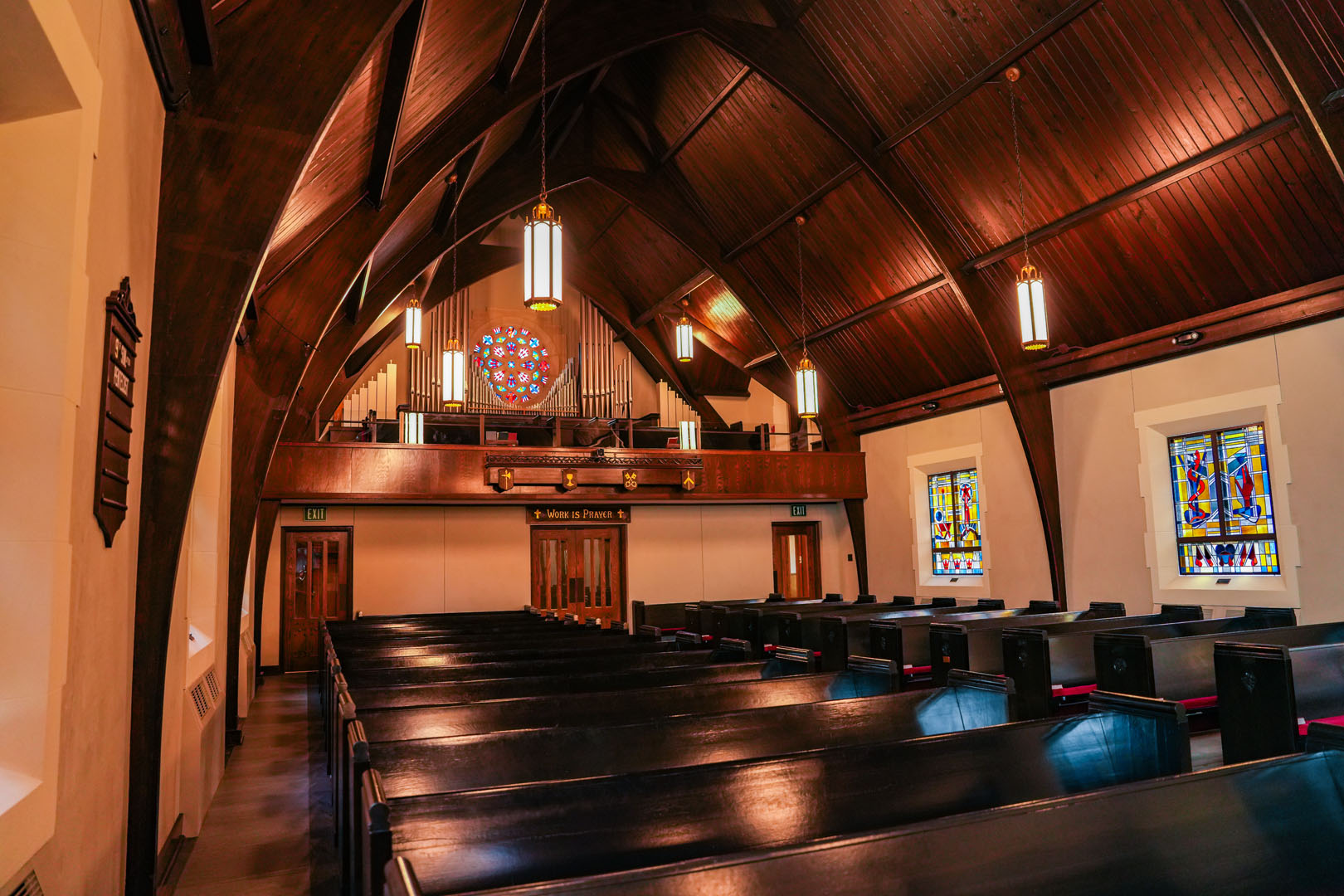 A well-lit interior of a church featuring wooden pews and a high, wooden arched ceiling. The back wall showcases a balcony with a pipe organ and a large round stained glass window. Additional stained glass windows line the side walls. Pendant lights hang from the ceiling.