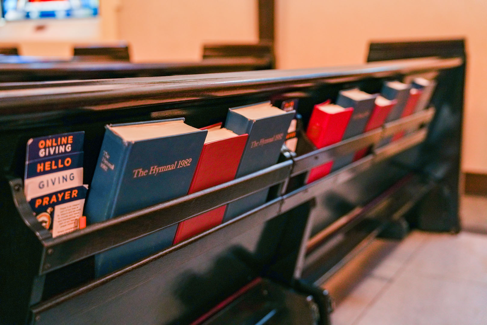 A church pew with several hymnals, prayer books, and pamphlets stored in the backrest pockets. The books are primarily blue and red, with one pamphlet promoting online giving visible from the side. The pews are made of dark wood.