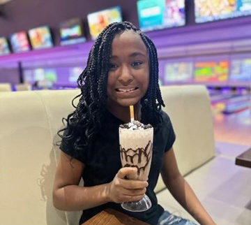 A young girl with braided hair sits in a booth at a restaurant or bowling alley, holding a tall glass of a chocolate milkshake topped with whipped cream and chocolate drizzle. Multiple colorful screens are visible in the blurred background.