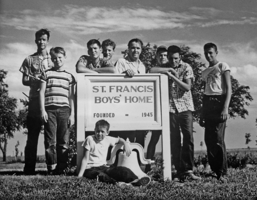 A black and white photo shows eight boys and one man posing around a sign that reads "St. Francis Boys' Home, Founded 1945." The group stands and kneels on a grassy area with trees in the background. Four boys are standing on the left, two boys and a man in the center, and two boys on the right. One boy is sitting on the ground in front.