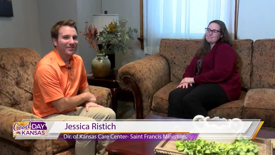 Two people are seated on brown sofas in a living room setting, engaged in conversation. The person on the left is wearing an orange shirt and beige pants, and the person on the right is wearing glasses, a black top, and maroon pants. A banner at the bottom reads "Good Day Kansas," and identifies the person on the right as "Jessica Ristich, Dir. of Kansas Care Center - Saint Francis Ministries.