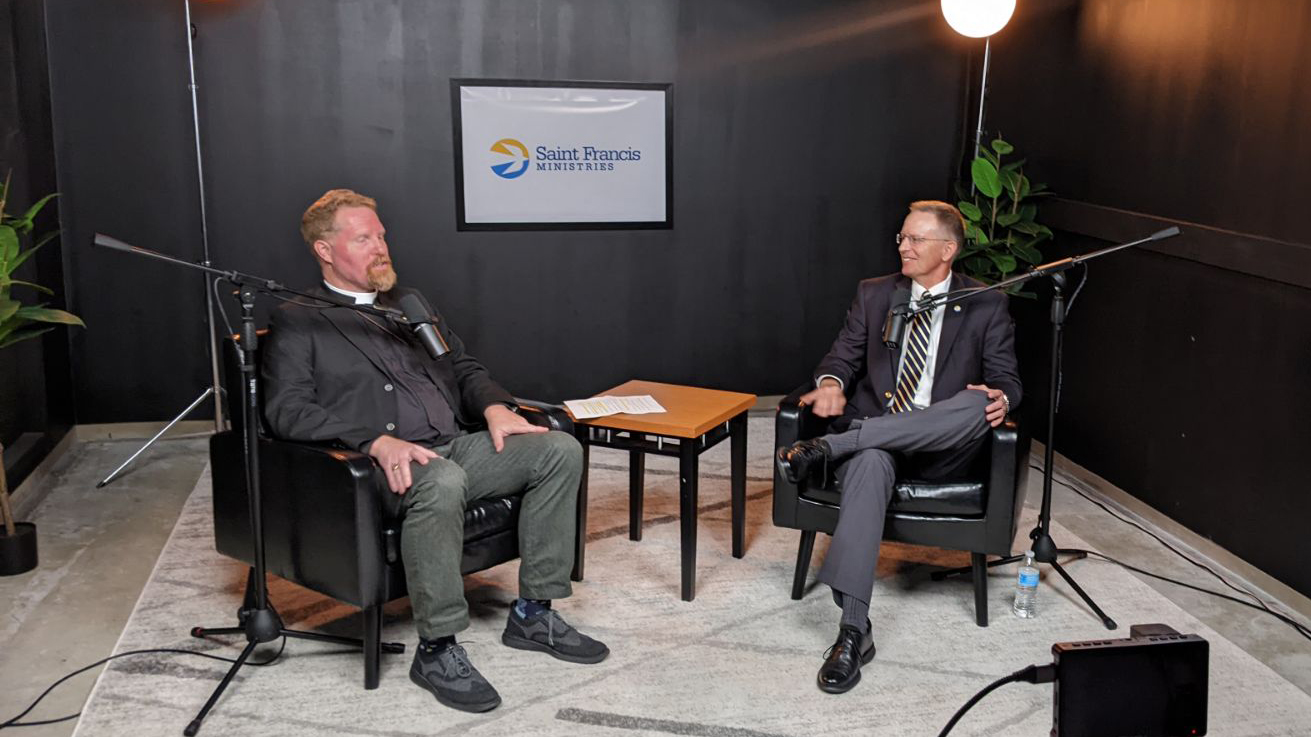 Two men are sitting and talking in a dimly lit room set up for an interview. One man has a microphone in front of him, and the other is facing him. The wall behind them features a logo that reads "Saint Francis Ministries." Two potted plants are placed on either side.