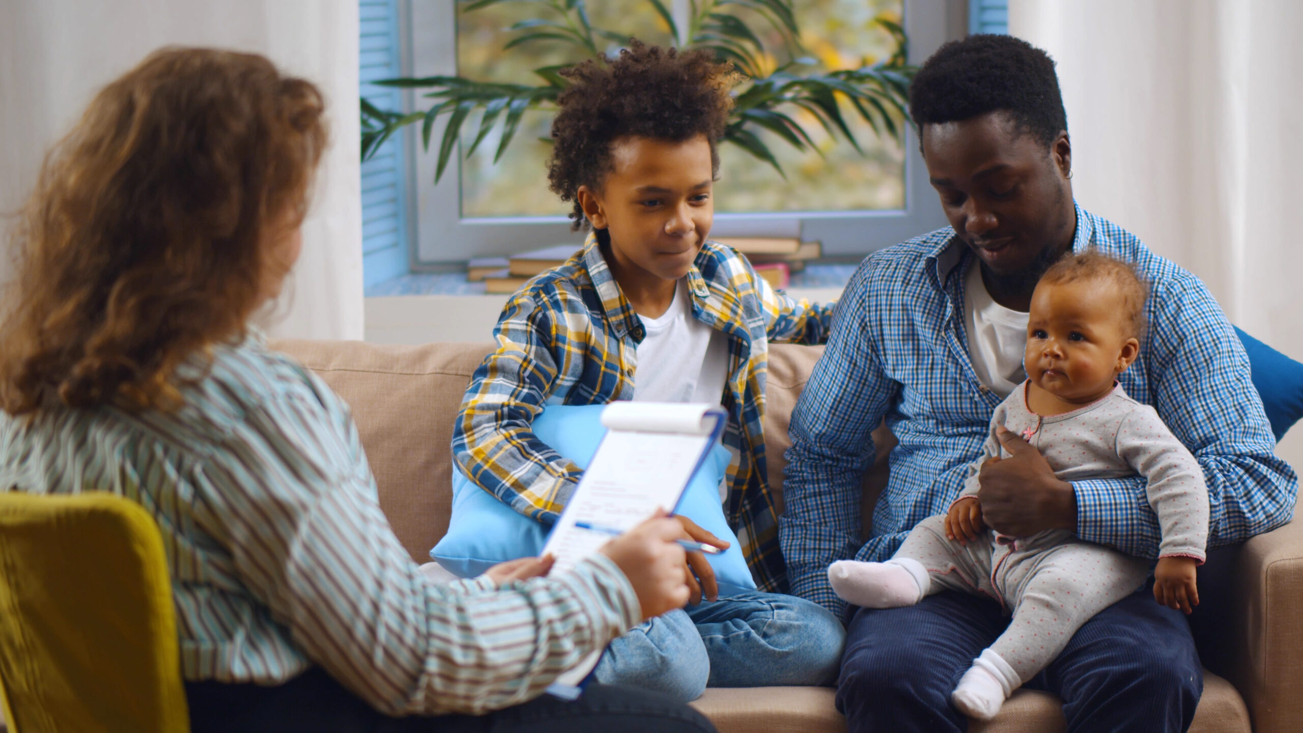 An adult woman with curly hair, holding a clipboard, is sitting on a yellow chair facing a man and two children. The man is holding a baby, and the older child is sitting on the sofa beside the man. They are in a well-lit room with a large window and plants.