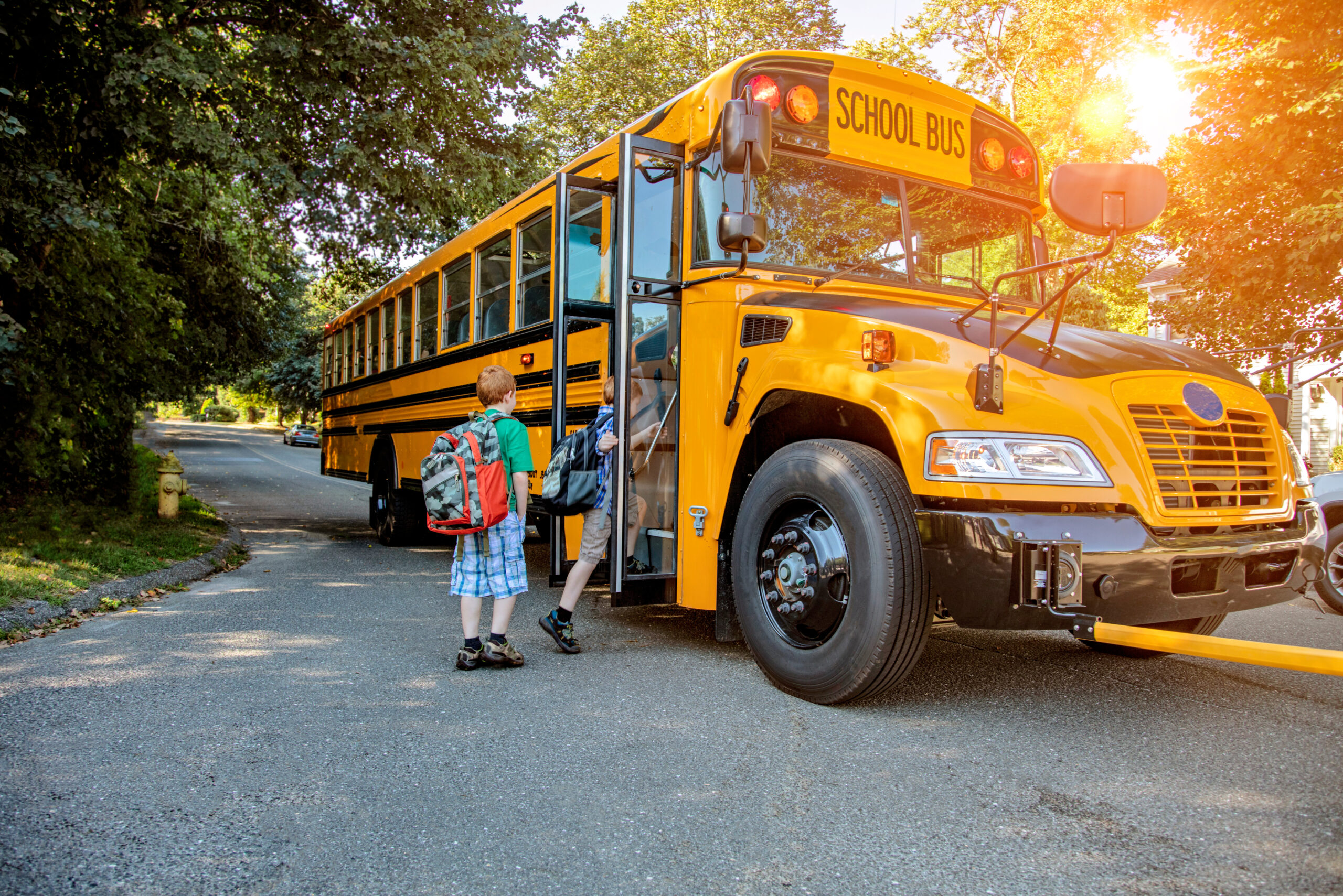 Two children with backpacks board a yellow school bus on a sunny day. The road is lined with trees, and the sun is shining through the leaves.