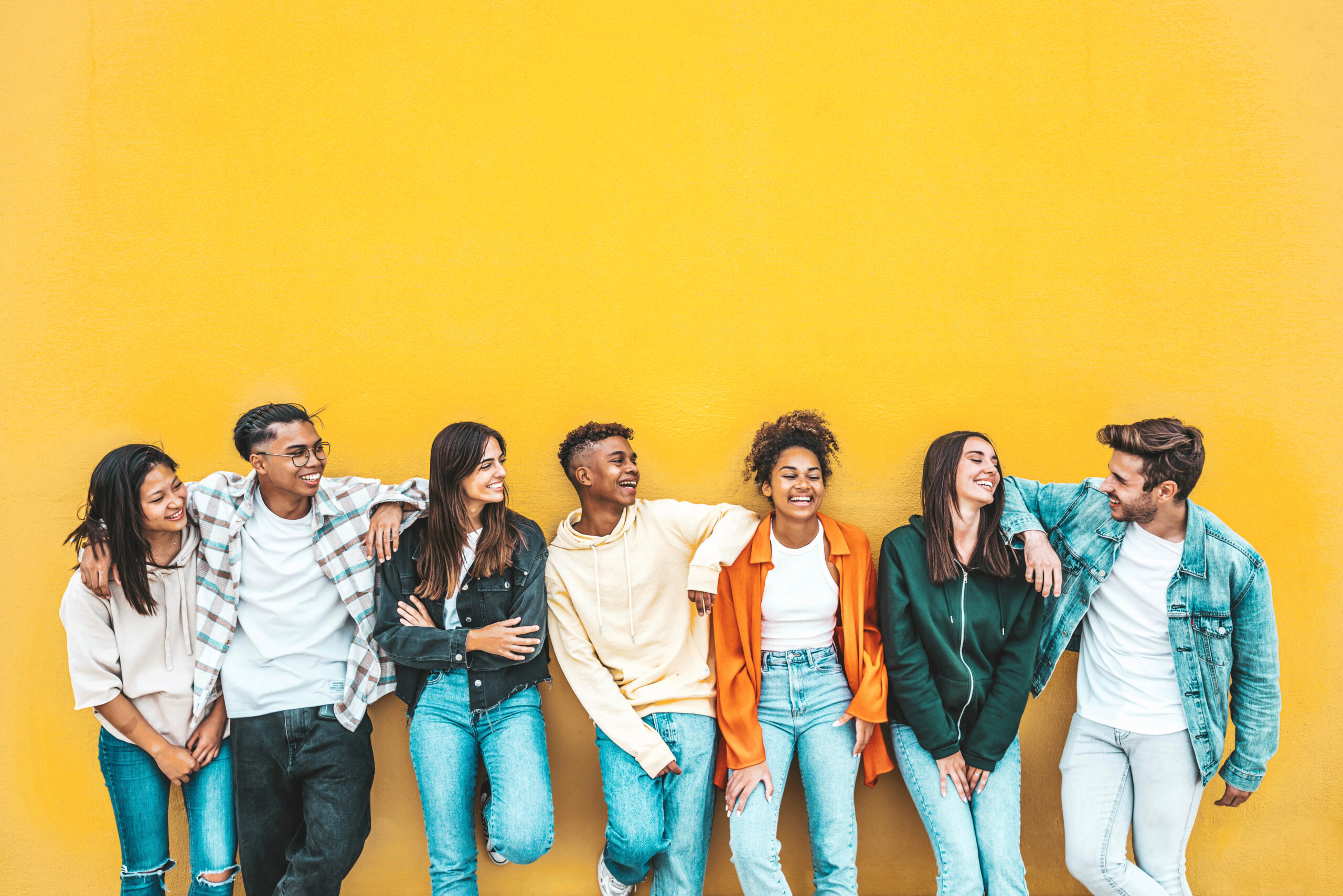 A group of seven young adults stand casually against a bright yellow wall. They are smiling and laughing, dressed in casual clothing, including jeans and jackets. The individuals have varied appearances and are interacting in a relaxed manner.