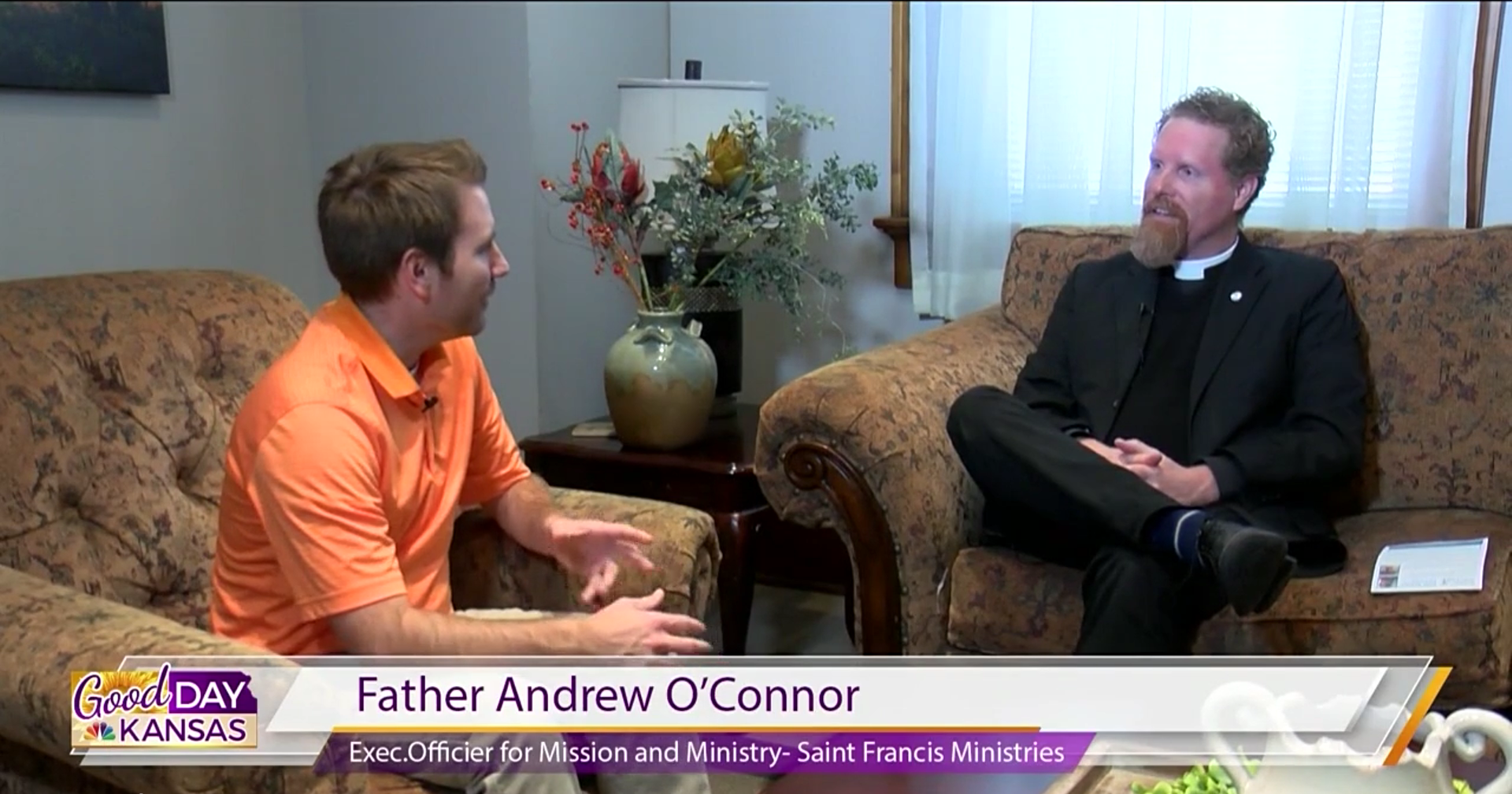 A person in an orange shirt interviews Father Andrew O'Connor, who is dressed in clerical attire, on a daytime talk show called "Good Day Kansas." They are seated in a cozy room with a floral arrangement and lamp visible in the background.