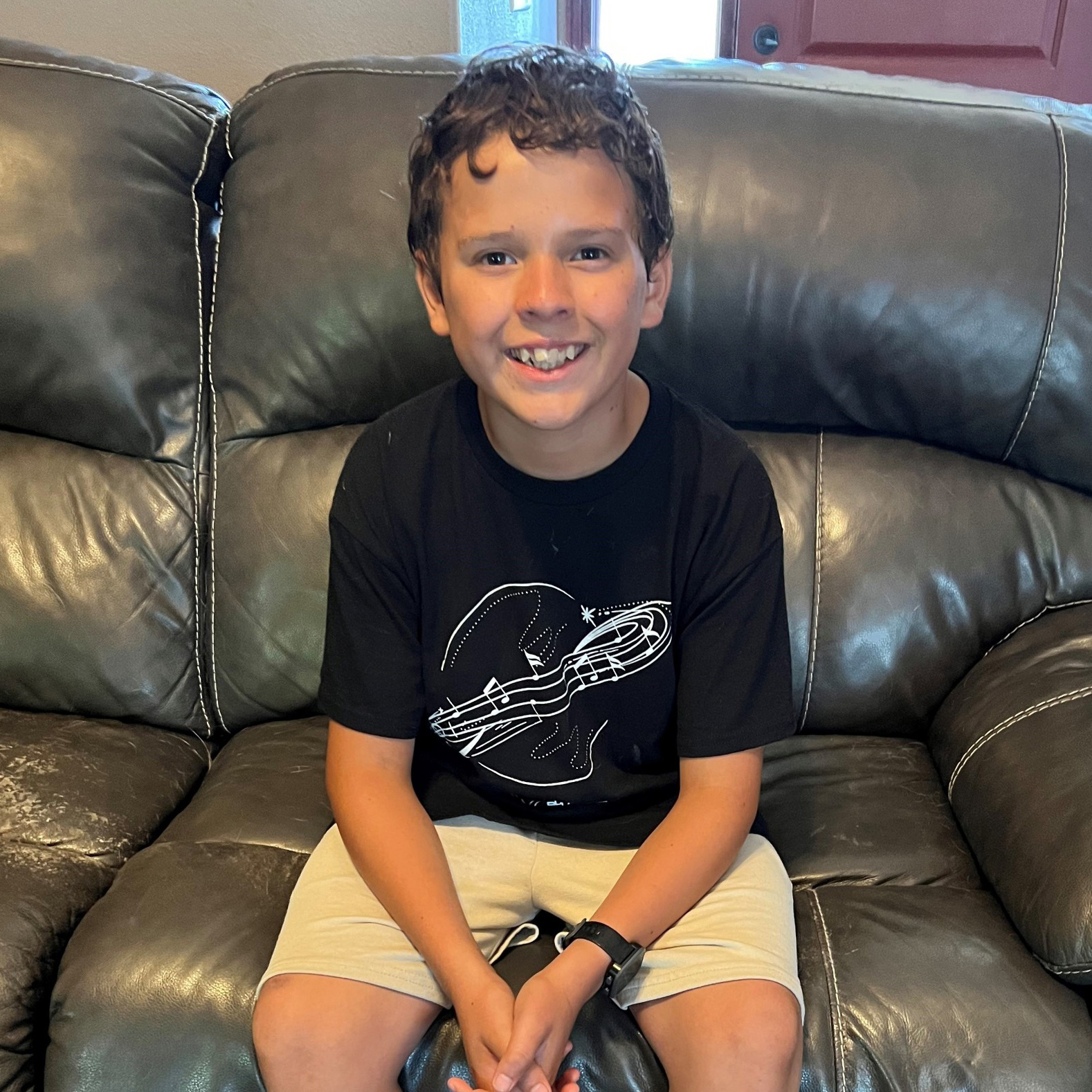 A young boy smiles while sitting on a black leather couch. He is wearing a black t-shirt with a white design featuring a planet and musical notes, along with beige shorts and a black wristwatch. The background includes part of a door and a beige wall.