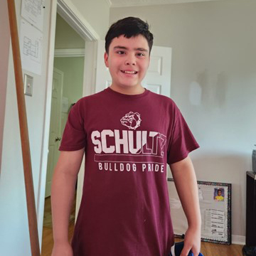 A young boy stands in a room wearing a maroon shirt that reads "SCHULTZ BULLDOG PRIDE." He is smiling and holding an object in his left hand. Behind him, a door is partially open and a picture frame rests against the wall to his right.